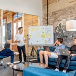 three men sitting while using laptops and watching man beside whiteboard